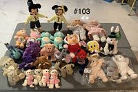 Full lot view showing all 27 plush dolls arranged on a grey surface, featuring Mickey Mouse dolls, Troll dolls, Care Bears, and others.