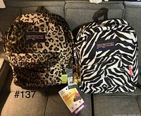 Front view of two JanSport backpacks side by side on a gray couch, one leopard print and one zebra print with tags attached.