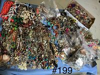 Wide view of mixed tangled scrap costume jewelry pieces piled with various colors, metals, and styles on a blue surface with boxes and plastic bags