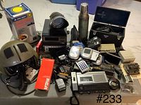 Photo showing assortment of vintage electronics, military helmet with cables, chargers, remote controls, and other small devices on a gray surface.
