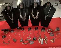 Photo showing multiple necklace and earring sets, and bracelets on black necklace stands and red surface with Paparazzi tags visible