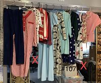 Wide shot of 13 vintage clothing pieces on hangers including pants, dresses, a blouse, and patterned garments in various colors.