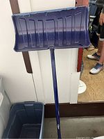 Blue plastic snow shovel with metal handle leaning against a wall.