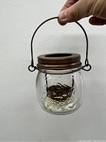 Photo of a clear glass hanging tealight candle holder with metal top rim and handle, held by a hand against a white wall.