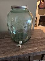 Full view of the large Mason jar style glass drink jug showing embossed glass lettering, metal screw lid, and metal spigot at the base.