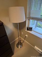 Photo of the IKEA dimmer floor lamp standing in a corner next to a window with blinds, plugged into a wall outlet, showing the full lamp and base.