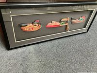 Long rectangular shadow box frame containing three Native carved art pieces mounted on grey fabric. Frame is black with glass front, measurements approximately 20x50 inches.