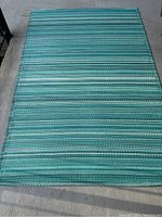 Photo of a rectangular outdoor patio rug with horizontal stripes in various shades of blue and green, lying flat on concrete.