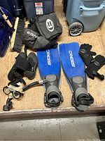 Photo showing the pair of blue Cressi fins, black diving gloves, and part of the mesh gear bag on a wooden floor.
