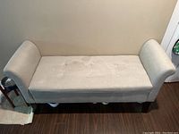 Front view of the light beige upholstered bench with tufted seat and rolled arms positioned against a wall on hardwood floor.