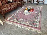 Rug lying flat on carpeted floor, showing floral pattern of pink roses and cream with fringe on edges.
