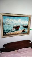 Full view of the framed oil painting hanging on a wall above a headboard, showing boats on shore and figures with a cloudy sky.