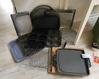 Full view of multiple metal bakeware pans including cookie sheets, trays, cooling racks, and non-stick skillet in box on floor