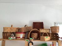 Wide view of 11 baskets on a wooden shelf showing a variety of styles including wood slat baskets, wicker baskets, and a painted strawberry basket.