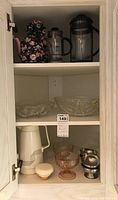 Cupboard with all items: floral teapot cozy with teapot, two glass French presses, clear glass bowls, two pink-toned glass pedestal bowls, white thermal carafe, and metal cups.
