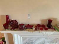 Full view of assorted coloured glassware items arranged on a white mantel with lace doilies. Includes vases, plates, candle holders, and decorative pieces mostly in shades of red.