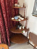 Wooden corner shelf with four triangular shelves against a wall with dark red floral curtains. Various small decorative items placed on shelves, not included in sale.