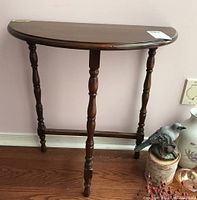 Wood demilune table with semi-circular top and four turned legs shown against a pink wall and hardwood floor, with a small bird figurine to the side.