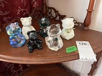 Six bear figurines displayed on a wooden surface: iridescent blue Fenton glass bear, white ceramic bear, black/greenish Fenton glass bear, clear glass bear, milky glass Fenton bear, and Oneida crystal bear.