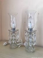Photo showing two matching glass lamps with etched hurricane shades and hanging glass prisms, cords coiled beside them.