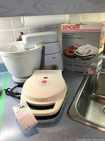 Singer waffle maker with box and Braun mixer displayed on kitchen counter near sink.