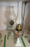 Two vintage oil lamps placed on a shelf. One lamp has a clear glass base with a frosted etched glass shade. The other lamp has a white glass and metal base with a clear glass chimney.