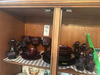 Cabinet shelf containing ruby glass items including stemware, plates, bowls, and cruets in deep red color.