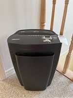 Full view of the black Fellowes shredder unit standing on carpet next to wooden stairs, showing the bin with handle and shredder top with slots.