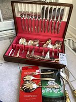 Open wooden box containing approximately 50 pieces of silver plated cutlery including knives, forks, spoons, serving spoons, and other serving pieces arranged inside the box with red lining. Polishing cloths and supplies positioned in front of the box.