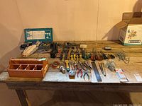 Photo of workshop bench top with wooden toolbox, pliers, wrenches, screwdrivers, tape measure, metal ruler, cutting knives, assorted fasteners and nails, peg board hooks, wire, and work gloves.