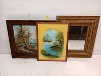 Two framed oil paintings and one ornate framed mirror with carved edges, all placed against a white background.