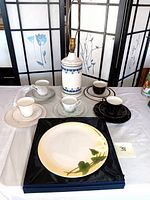 Five sets of teacup, saucer, and plate with various patterns including black and gold, white floral, and blue botanical, arranged with a blue on white ceramic lamp in center and a boxed calla lily plate on black fabric.