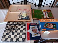 Chess board and game sets displayed on a table, including marble-style chess board, baseball player figures in a plastic bag inside the chess case, Shut the Box game, and oversized playing cards.