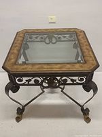 Front view of the square center table showing the glass inset, brown frame, ornate wrought iron base with scroll detail and golden claw feet.