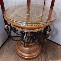 Front angled view of the ornate table showing the wood base, carved sunflower center, metal arms, and glass top.