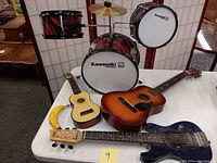 Full set of toddler drum kit, acoustic guitar, electric guitar, and ukulele on a white table