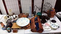 Overview of miscellaneous household items including decorative plates, plant holders, a wicker wine basket, clay pots, and more on a white table.