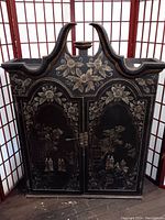 Full front view of vintage Asian hutch top, showing black hand painted finish with floral and scenic motifs, two cabinet doors with metal latch, curved pagoda style top with central holder, scratches on surface.