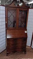 Front view of vintage wooden secretary desk with glass doors, drop-front writing surface, and curved drawers.
