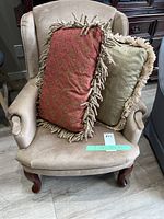 Full view of beige armchair with two decorative fringed cushions, showing shape, upholstery, and legs.