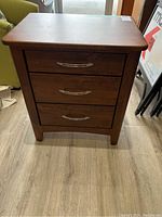 Front view of wooden bedside table showing three drawers and silver handles, standing on a wood plank floor near a green couch.