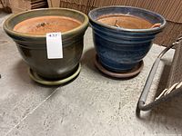 Two ceramic pots side by side on a concrete floor, green and blue respectively, with drainage saucers underneath.