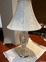 Full view of lamp showing fabric lampshade and glass base on chrome platform stand with attached power cord and plug.