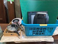 Photo showing Honeywell heater inside blue laundry basket along with a framed picture behind it. Kitchen pans and cutlery tray positioned next to basket.