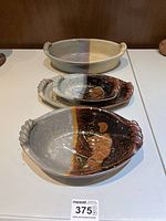 Four oval ceramic serving dishes stacked, showing dual-tone glaze with natural beige and dark brown, and braided handles on each end.