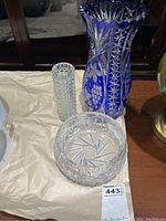 Three items on beige surface: tall blue glass vase with cut pattern, smaller clear textured glass vase, and clear crystal serving bowl with cut pattern