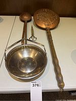 Vintage copper bed warmer, two long handled copper ladles, vintage metal scissors with brass finish, and a polished metal serving bowl with handles on a white table.