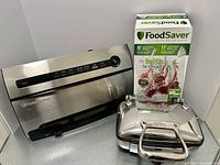 FoodSaver V3825 vacuum sealer beside box of vacuum sealing rolls and bags, alongside All-Clad waffle maker.