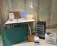 Full view of drafting table with art supplies, lamp, storage carts, cutting mats, and crafting tools setup in a basement room with concrete walls