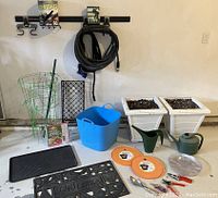 Photo of the lot showing garden hoses coiled on wall rack, two green tomato cages, black plastic boot tray, two white planters filled with soil, blue plastic bin, two green watering cans, black doormat with welcome text, secateurs, spade, and other accessories.